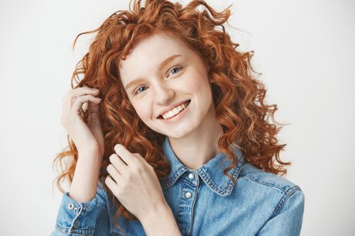A girl with long, curly red hair wearing a denim shirt, smiling at the camera against a white background