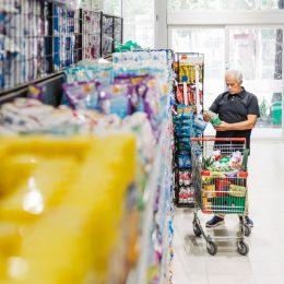 Senior man with shopping cart looking at grocery products in supermarket aisle. Mature male customer looking at groceries in store.