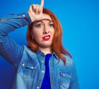 woman doing the "loser" sign on her forehead against a blue background