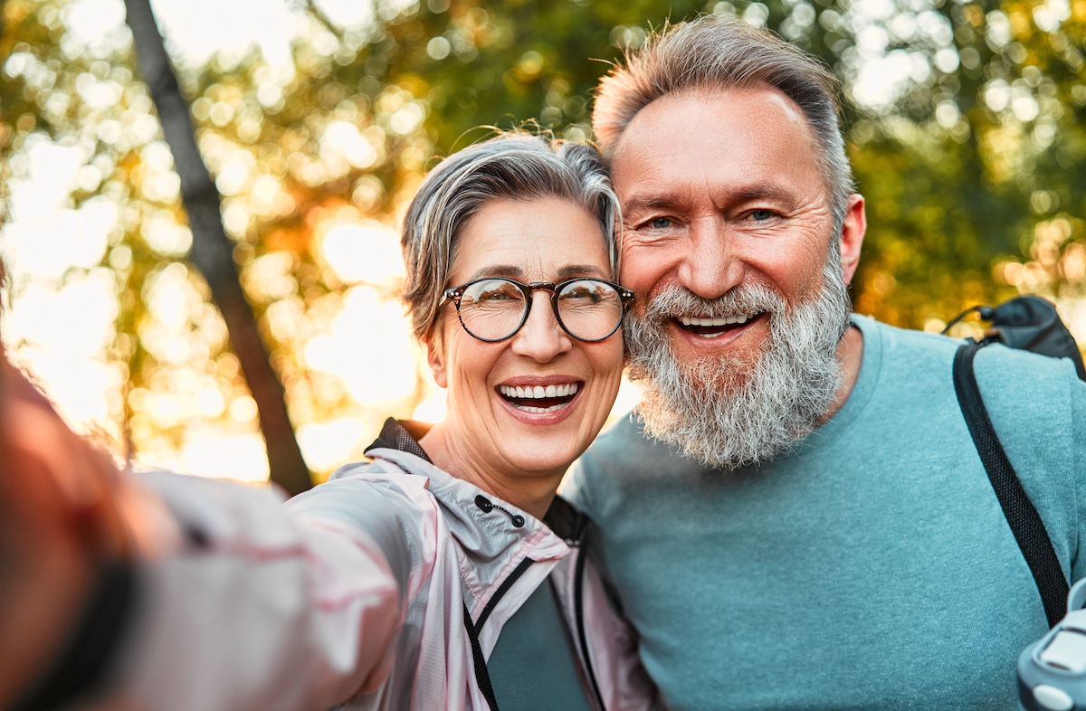A happy, mature couple both with gray hair take a smiling selfie while standing outside among trees