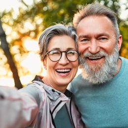 A happy, mature couple both with gray hair take a smiling selfie while standing outside among trees