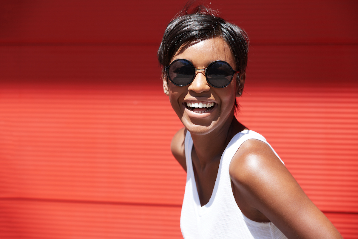 A smiling woman with a pixie haircut wearing sunglasses and a white tank top against a red background