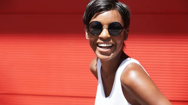 A smiling woman with a pixie haircut wearing sunglasses and a white tank top against a red background