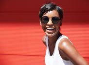 A smiling woman with a pixie haircut wearing sunglasses and a white tank top against a red background
