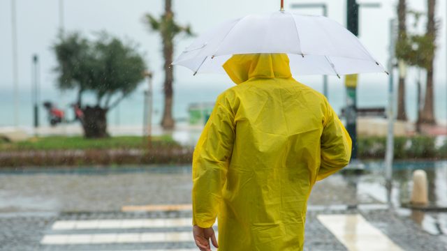 A man standing in a crosswalk in a rainstorm while wearing a raincoat and using an umbrella