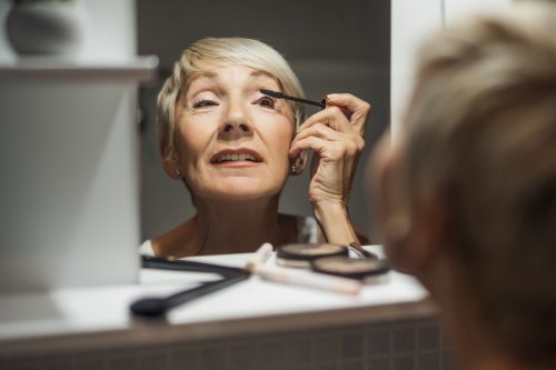 woman with short blonde hair applying mascara in the bathroom mirror
