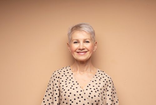 Mature smiling woman with a short pixie haircut wearing a beige dress with black polka dots against a beige background