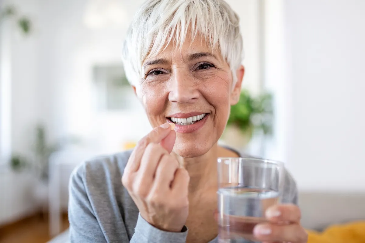 A happy mature woman with short white hair wearing a gray long-sleeved shirt takes a vitamin with a glass of water