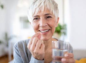 A happy mature woman with short white hair wearing a gray long-sleeved shirt takes a vitamin with a glass of water