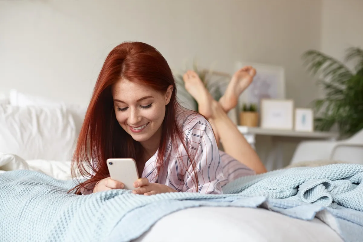 woman with red hair laying on her stomach in bed, using her phone