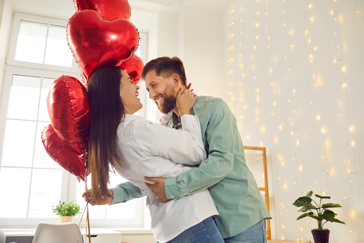 A happy, romantic couple hugging in their living room; the man is holding red heart-shaped balloons