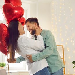 A happy, romantic couple hugging in their living room; the man is holding red heart-shaped balloons
