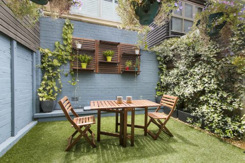 Small backyard surrounded by gray brick walls with climbing ivy. There's a small wood dining set and hanging planters.