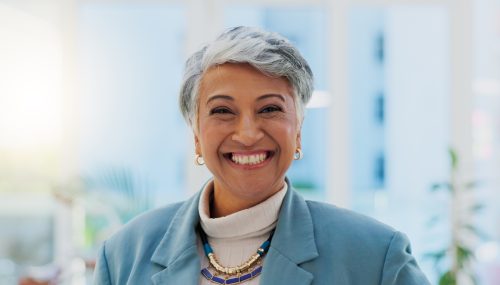 Portrait of a mature woman with short gray hair smiling big in her office while wearing a blue blazer and white turtleneck