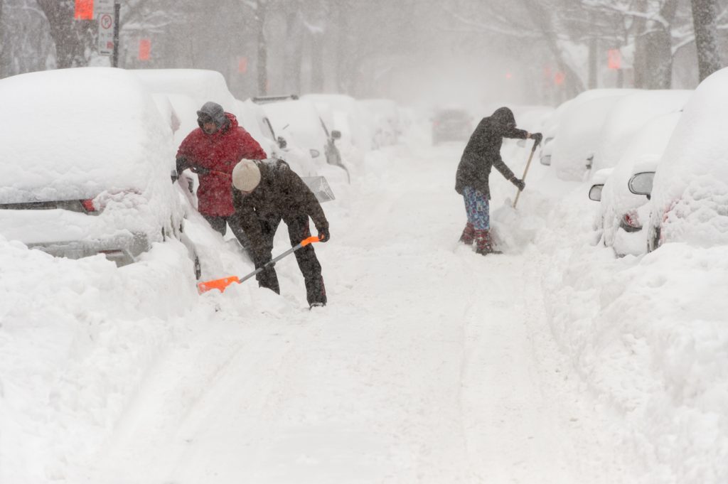 Three people shoveling out cars on a street during a heavy snowstorm