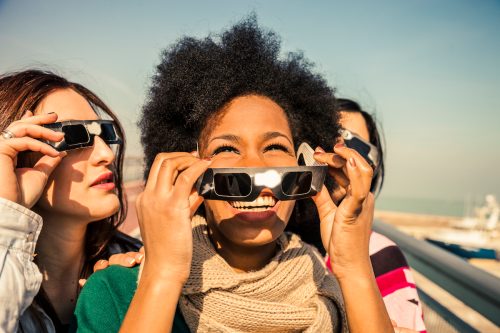 Three happy female friends looking at a solar eclipse, as one woman removes her eclipse glasses