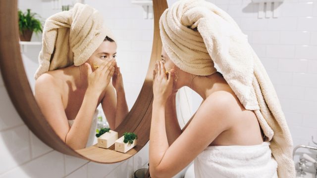 Young happy woman in towel applying organic face scrub and looking at round mirror in stylish bathroom. Girl making facial massage, peeling and cleaning skin on face. Skin Care and Hygiene