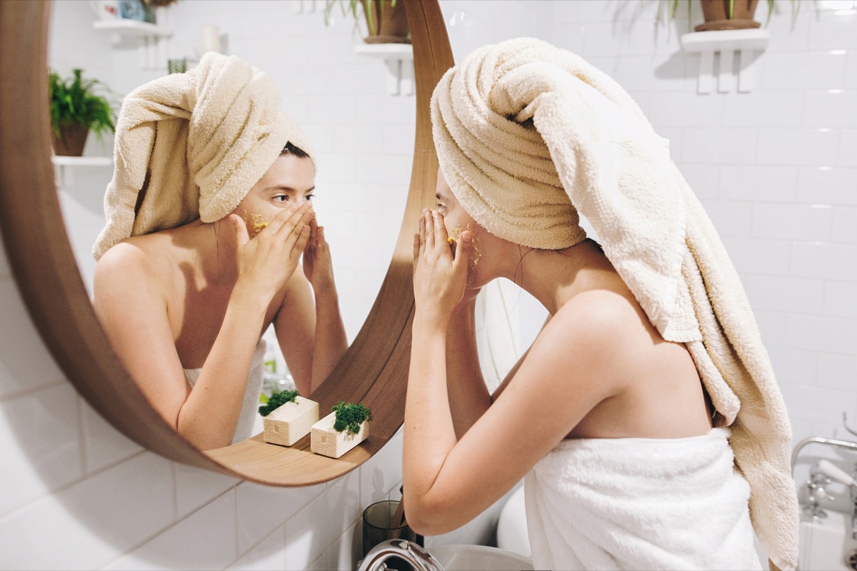 Young happy woman in towel applying face mask while looking in round mirror in bathroom. 