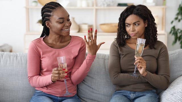 Happy woman showing her upset and jealous girlfriend her engagement ring while they sip champagne on the couch.