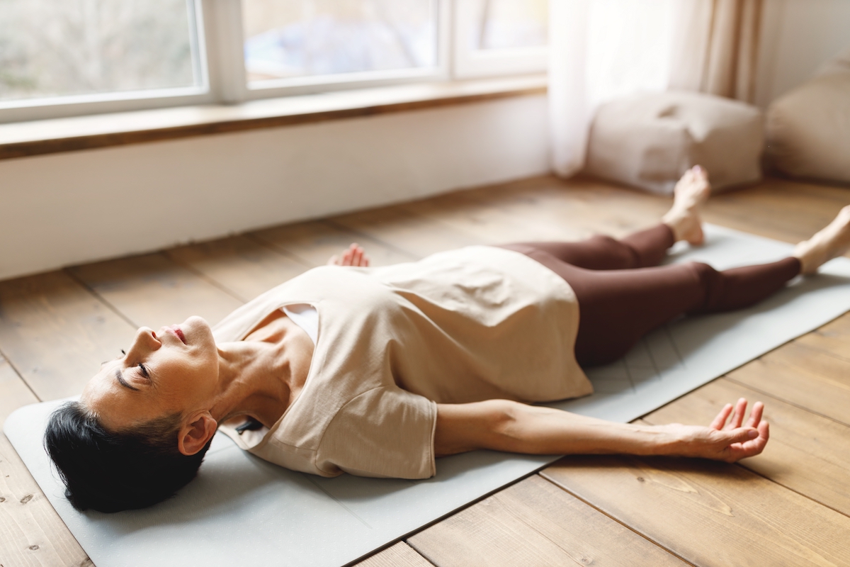 A woman wearing activewear lays on her back on her yoga mat in Shavasana pose