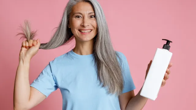 A smiling mature woman with long gray hair, wearing a light-blue t-shirt, holds her hair in one hand and a blank shampoo bottle in the other, against a light-pink background