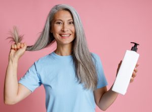 A smiling mature woman with long gray hair, wearing a light-blue t-shirt, holds her hair in one hand and a blank shampoo bottle in the other, against a light-pink background