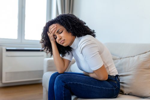 A woman sitting on the couch and holding her stomach and head in pain