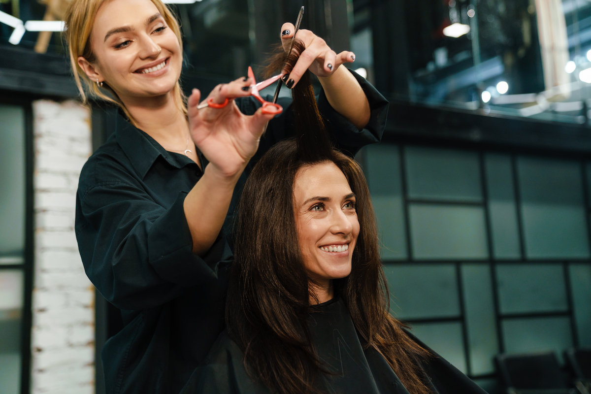 A young, smiling blonde woman cutting the long, dark hair of her client.