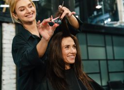 A young, smiling blonde woman cutting the long, dark hair of her client.