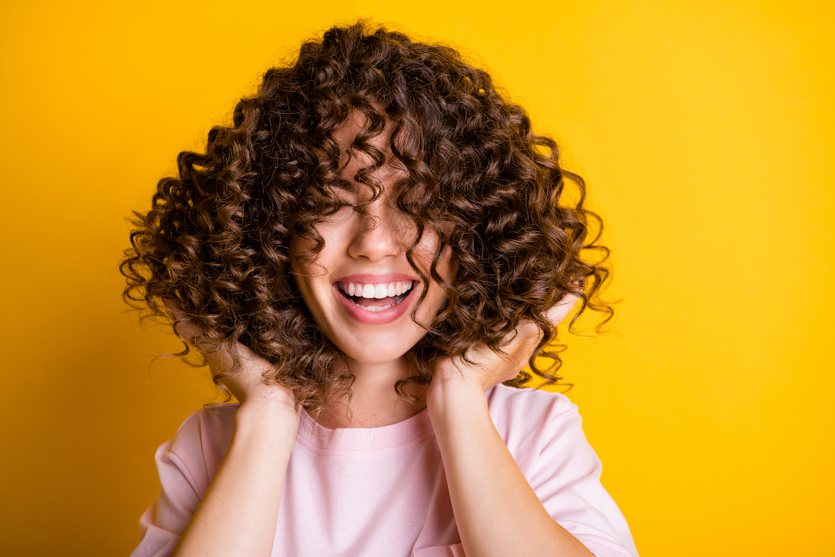 Portrait of a young woman with curly brown hair wearing a pink t-shirt laughing and touching her hair isolated on bright yellow background