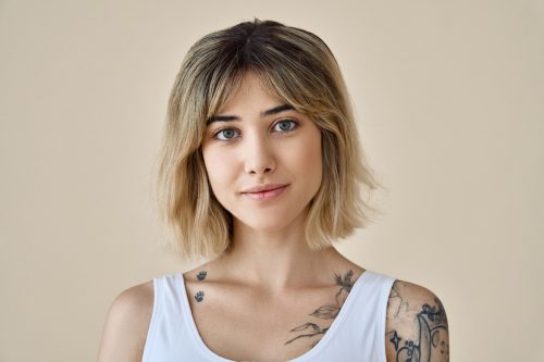 Portrait of a young woman with a short blonde shag haircut and tattoos wearing a white tank top against a beige background