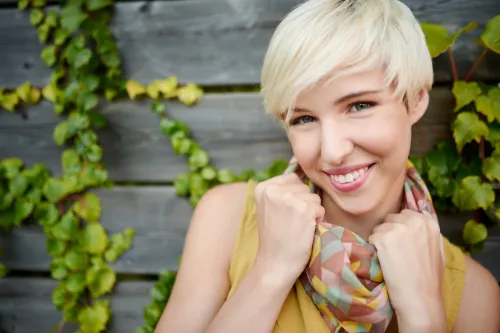 Portrait of a young woman with a platinum blonde pixie haircut standing against a wood wall with ivy