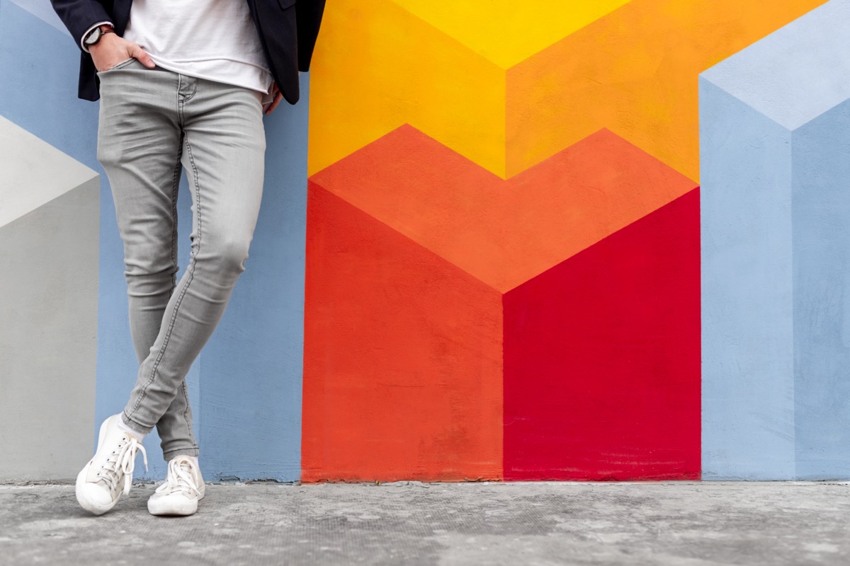Crop anonymous male in trendy gray skinny jeans and white sneakers holding hand in pocket while leaning against colorful wall