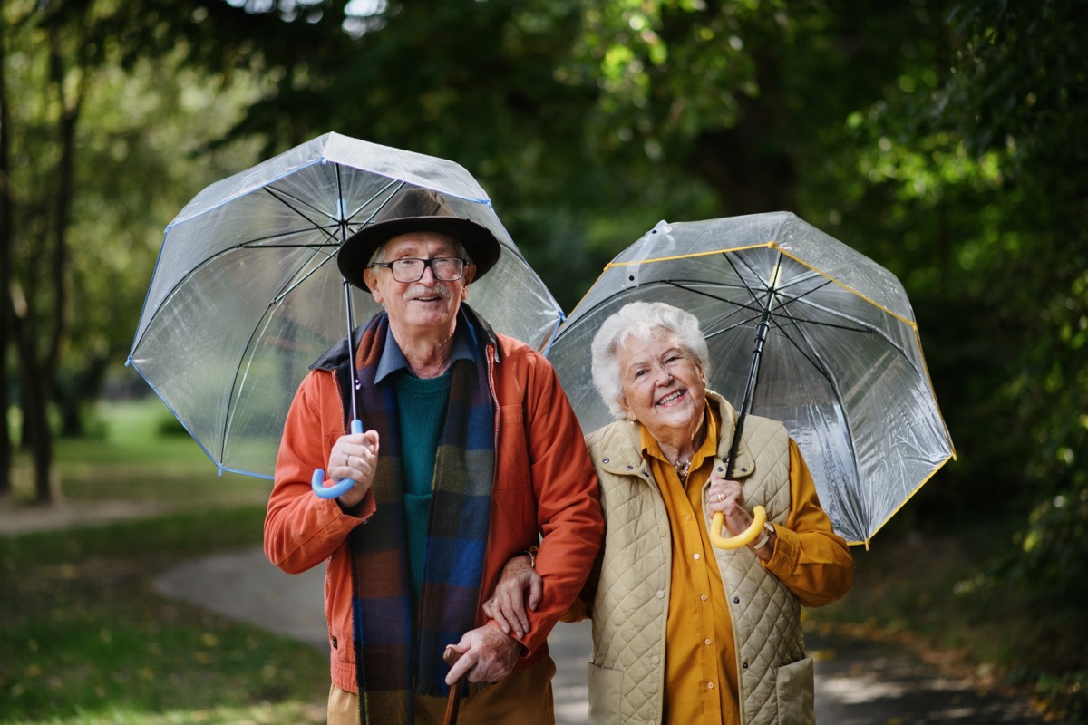 Happy senior couple walking with umbrellas in city park together.