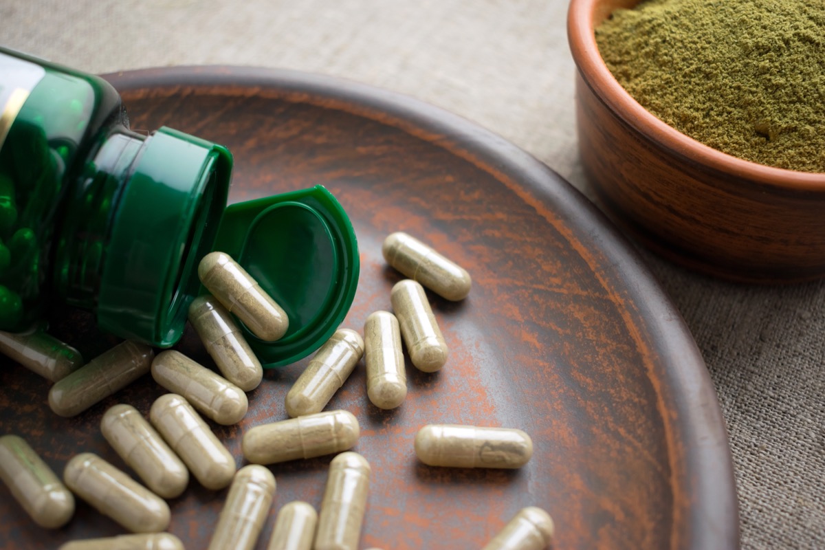 Green tea supplements spilling out of bottle onto wood bowl with a bowl of green tea powder in the background