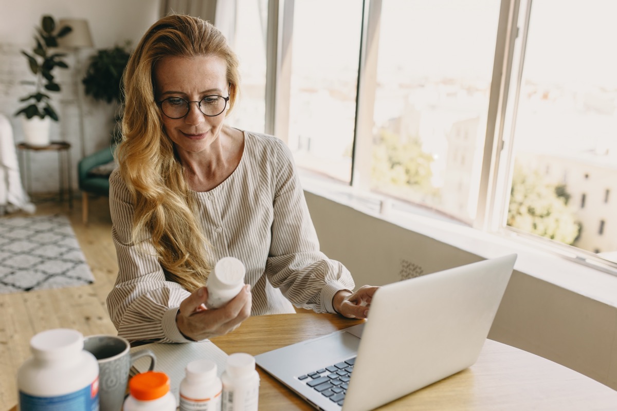 Mature woman sitting in her home at her laptop looking at the label of a supplement bottle
