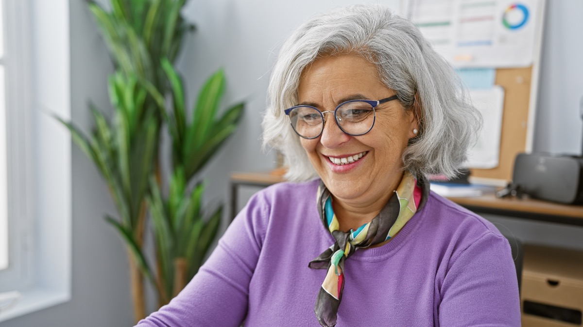Smiling mature woman with gray hair in an office, wearing a purple sweater