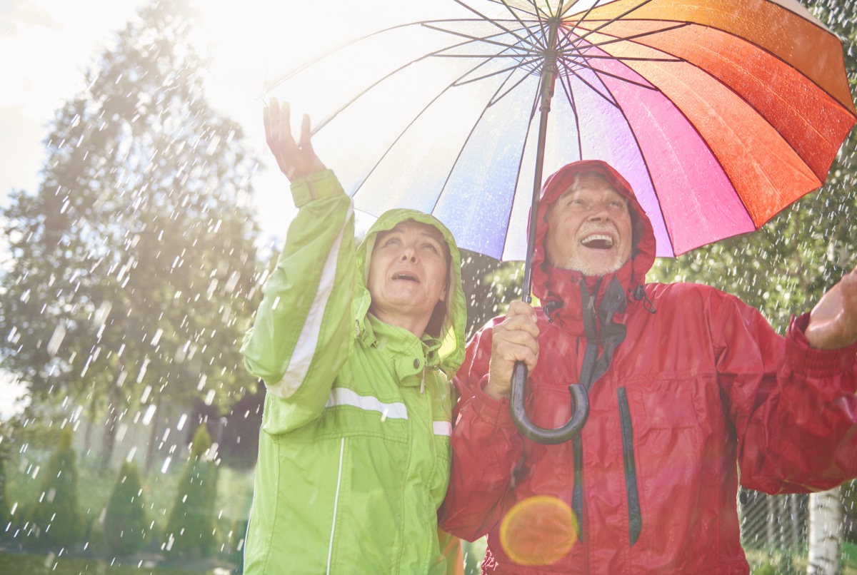 Senior couple wearing green and red raincoats holding a rainbow umbrella playing in the rain