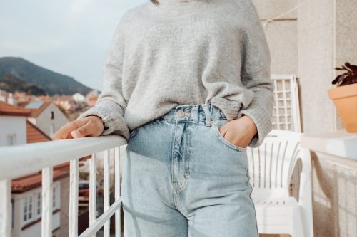 Close up of a woman wearing mom jeans and a gray sweater while standing on a balcony