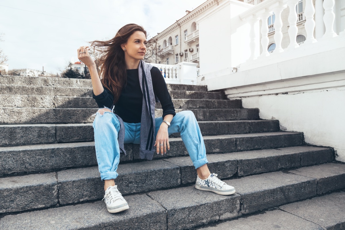 Woman wearing a black shirt, ripped boyfriend jeans and sneakers posing on stairs outside in an old city