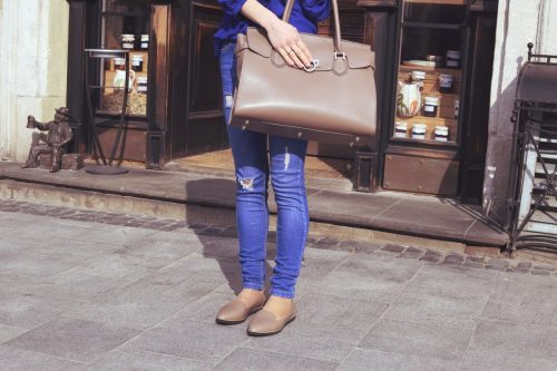 Young stylish girl in blue jeans with holes with big light brown handbag in hands near the shop in the city. Summer or spring stylish outfit, street fashion look. Woman with handbag
