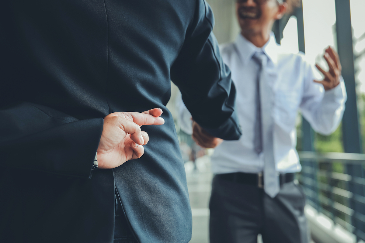 Man in a suit crossing his fingers behind his back while shaking hands with a male coworker