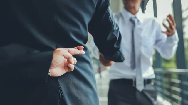 Man in a suit crossing his fingers behind his back while shaking hands with a male coworker