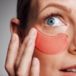 Closeup of a woman applying a peach under eye patch against a gray background