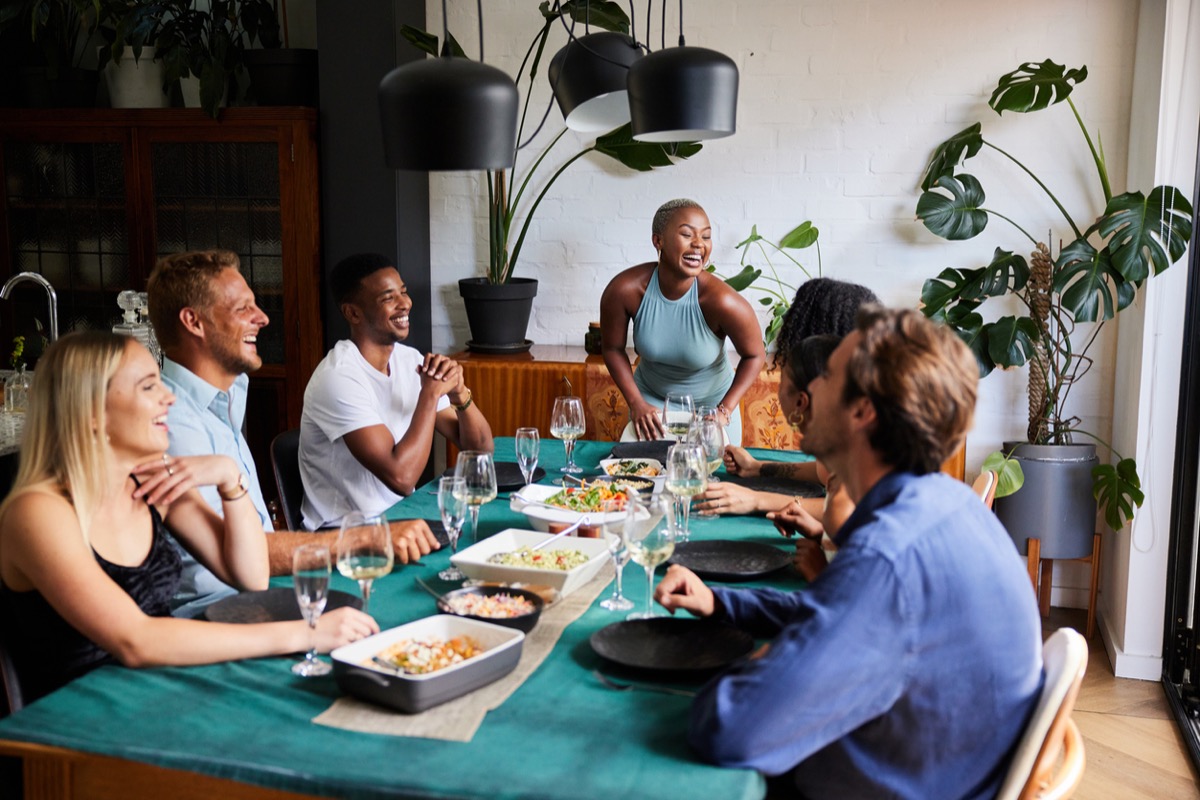 Young woman laughing at the head of a table while hosting a dinner party for a diverse group of young friends at her home