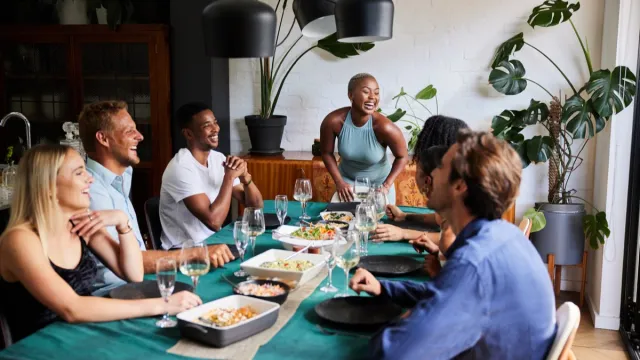 Young woman laughing at the head of a table while hosting a dinner party for a diverse group of young friends at her home