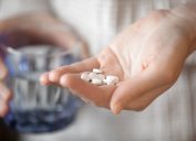 Healthcare, treatment, supplements concept photo. Woman arm holding heap of small round meds and glass of water before taking medication, shallow depth of field, focus on medicine