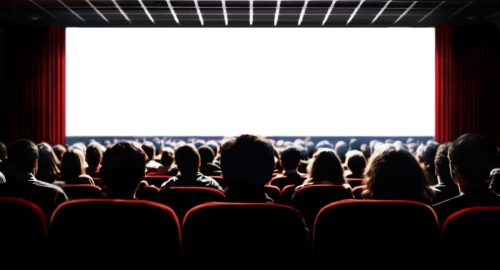 Movie theater audience looking at white screen, pictured from behind