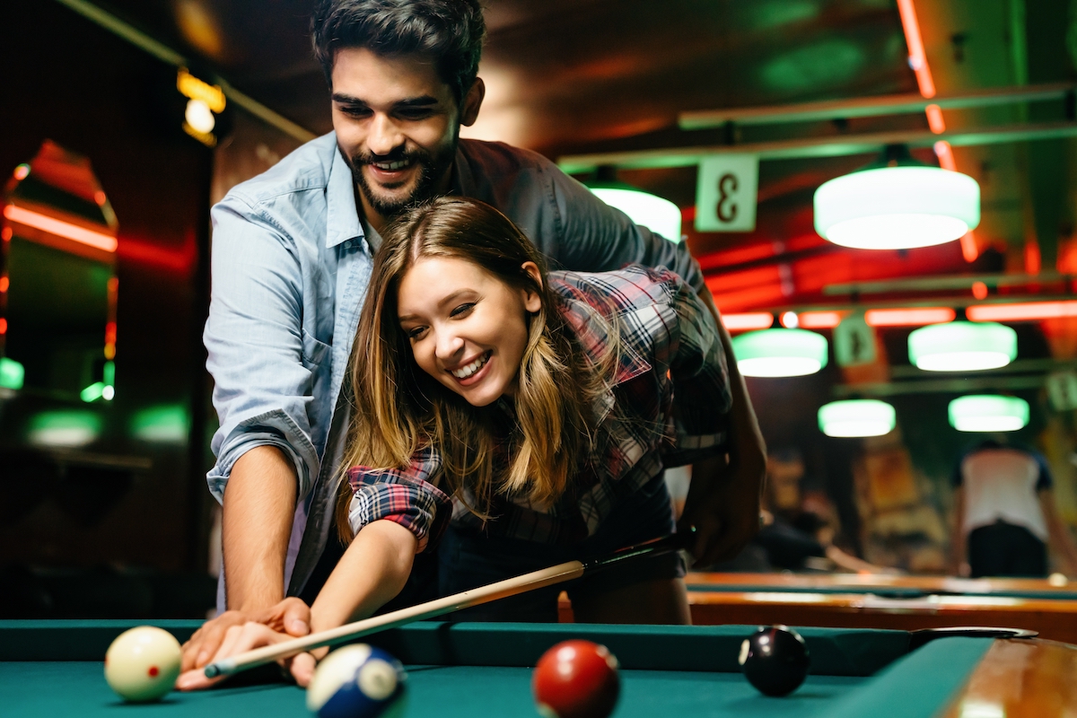 Portrait of young couple having fun playing billiard together. The man is showing the woman how to shoot pool.