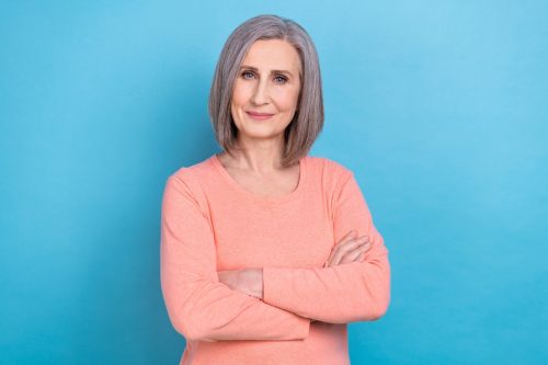 Portrait of a mature woman with gray hair wearing a peach shirt standing against a light-blue background
